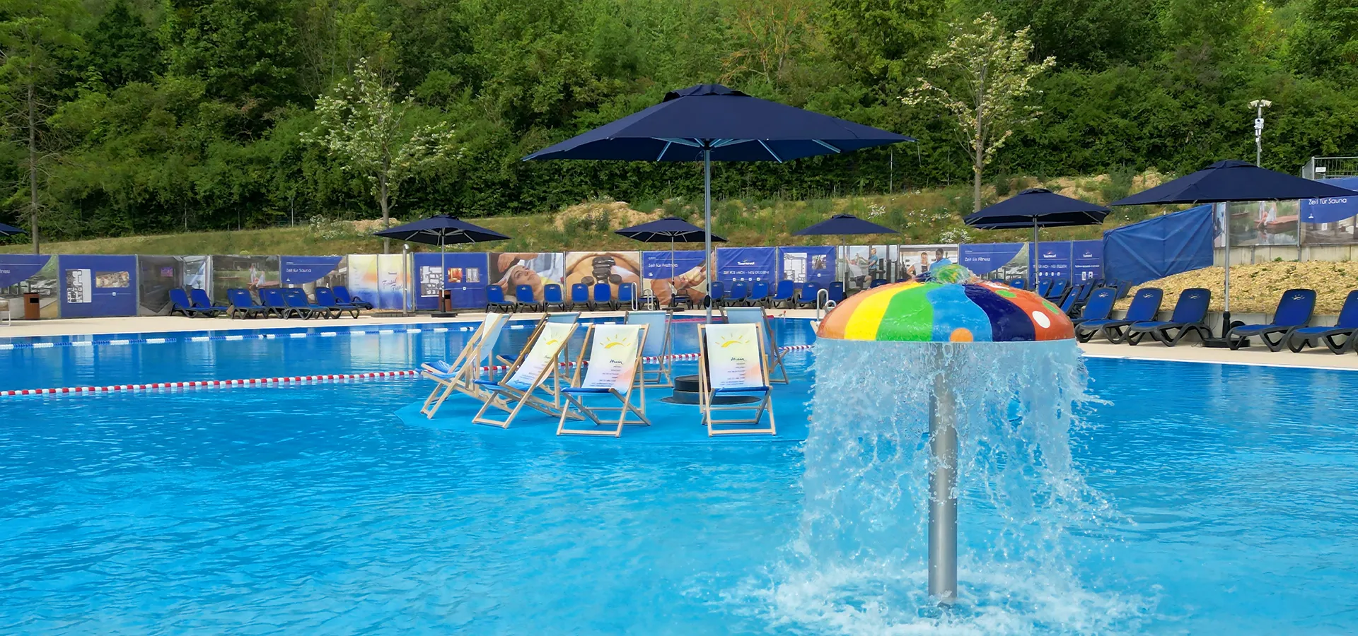 Blick über die Wasserfläche mit Wasserpilz und Liegestühlen