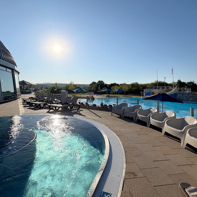 Sonniger Außenbereich des Freizeitbeckens mit geschwungenem Beckenrand. Entlang der Terrasse stehen weiße Loungesessel und Sonnenliegen. Im Hintergrund erstreckt sich das große Freibad mit blauen Sonnenschirmen und einer Wasserrutsche.