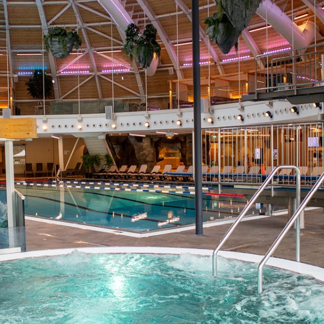 Blick vom sprudelnden Whirlpool auf das Sportbecken mit abgetrennten Schwimmbahnen und Sprunganlage mit 1-Meter- und 3-Meter-Sprungbrett. Die Halle verfügt über eine hohe Holzkuppel, farbige Beleuchtung, hängende Pflanzendekorationen und Ruheliegen am Beckenrand.