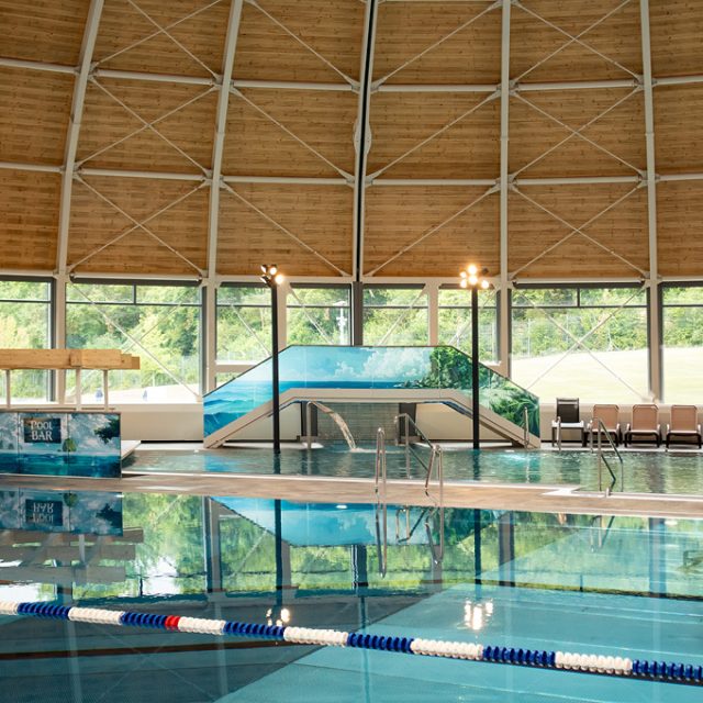 Großzügiges Hallenbad mit türkisblauem Schwimmbecken, hoher gewölbter Holzdecke und großen Panoramafenstern mit Blick ins Grüne. Im Hintergrund sind eine Poolbar, eine kleine Brücke über dem Wasser und mehrere Ruheliegen zu sehen.