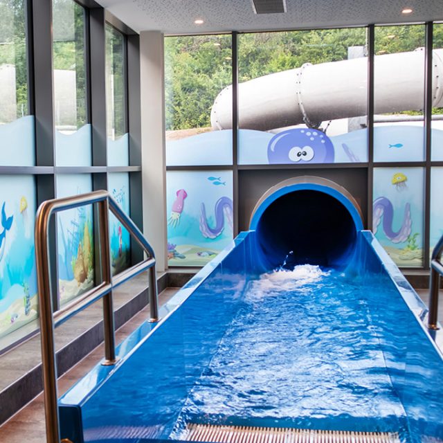 Auslauf der Röhrenrutsche „Blaue Krakenrutsche“ mit blauem Landebecken und seitlichen Edelstahlgeländern. Große Fenster mit bunten Unterwassermotiven zeigen Fische, Meerespflanzen und einen Kraken. Draußen ist die weiße Röhrenrutsche zu sehen.