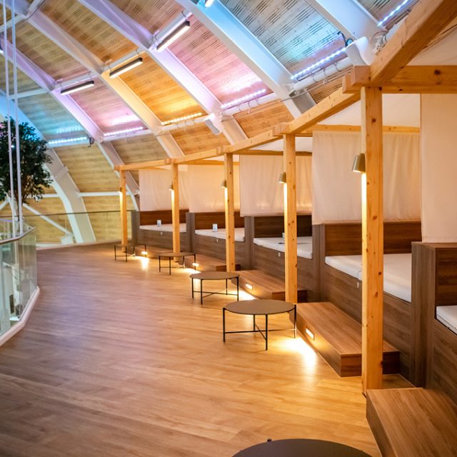 Panorama-Cabanas mit weißen Liegeflächen, Vorhängen und Holzrahmen auf der Empore des Schwimmbads. Warme Beleuchtung und kleine Beistelltische sorgen für eine gemütliche Atmosphäre. Eine Glasbrüstung ermöglicht den Blick auf die darunterliegende Badelandschaft.