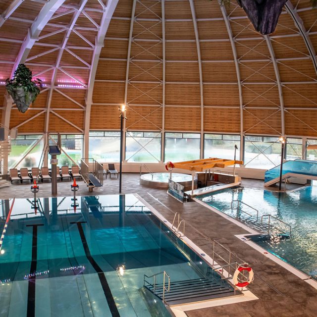 Helles Hallenbad mit Schwimmbecken, Freizeitbecken, Poolbar und hoher, gewölbter Holzdecke. Große Fenster bieten einen Blick auf den Außenbereich.